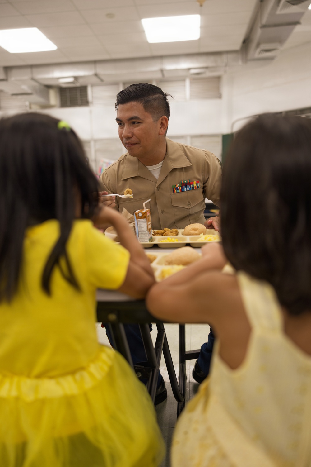 A military service member in tan/khaki uniform with service ribbons is seated at a cafeteria table sharing a meal with two young children dressed in yellow. The setting appears to be a school or institutional cafeteria with fluorescent lighting and standard cafeteria trays. The interaction appears warm and social, suggesting a community outreach or family visit event.