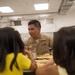 A military service member in tan/khaki uniform with service ribbons is seated at a cafeteria table sharing a meal with two young children dressed in yellow. The setting appears to be a school or institutional cafeteria with fluorescent lighting and standard cafeteria trays. The interaction appears warm and social, suggesting a community outreach or family visit event.