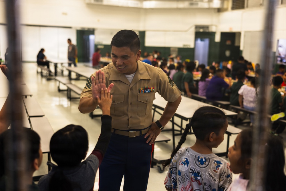 A U.S. Marine in service uniform is giving a high-five to a child in what appears to be a school cafeteria or gymnasium. The Marine is smiling warmly while interacting with several children in the foreground. The background shows a large group of people, including children and adults, seated at long cafeteria-style tables.