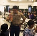 A U.S. Marine in service uniform is giving a high-five to a child in what appears to be a school cafeteria or gymnasium. The Marine is smiling warmly while interacting with several children in the foreground. The background shows a large group of people, including children and adults, seated at long cafeteria-style tables.