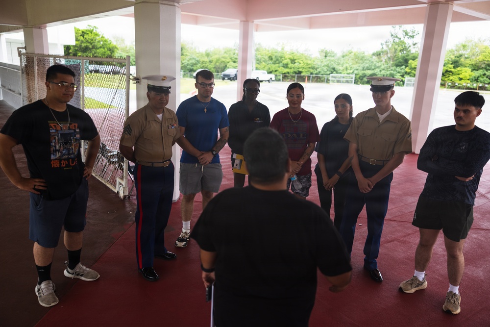 A group of approximately nine individuals, including at least two uniformed U.S. Marines in service uniforms with white covers, are gathered in a semi-circle under a covered outdoor structure. A person with their back to the camera appears to be addressing or briefing the group. The setting appears to be a military or institutional facility with a red-painted floor and white columns.