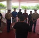 A group of approximately nine individuals, including at least two uniformed U.S. Marines in service uniforms with white covers, are gathered in a semi-circle under a covered outdoor structure. A person with their back to the camera appears to be addressing or briefing the group. The setting appears to be a military or institutional facility with a red-painted floor and white columns.