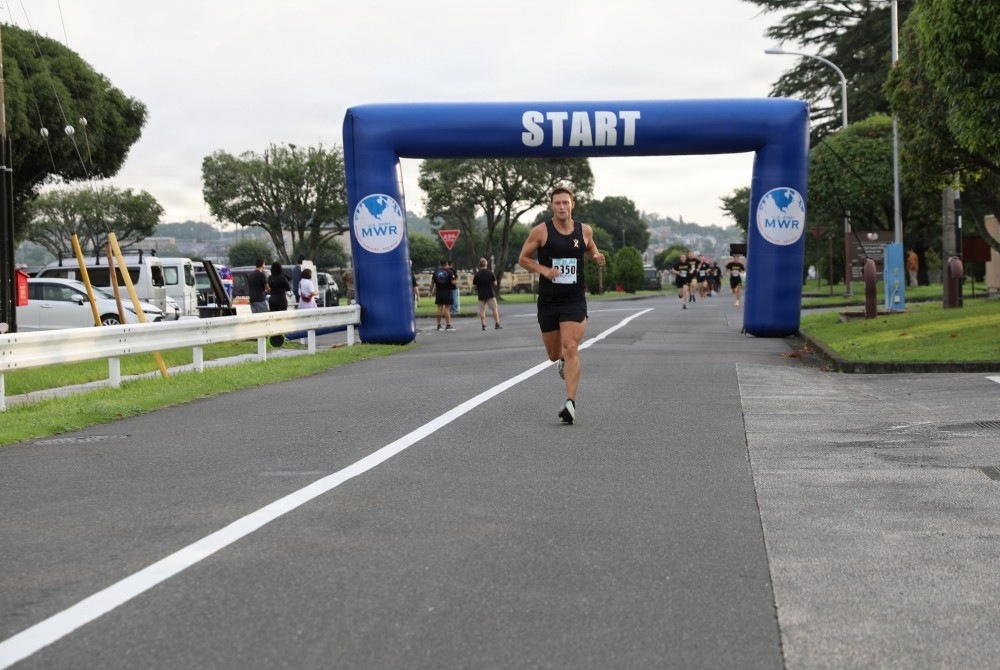 A running race event is underway on a paved road, with a prominent blue inflatable 'START' arch sponsored by MWR (Morale, Welfare and Recreation). A male runner wearing bib number 2350 and a black tank top is the lead runner passing under the arch. Additional runners and spectators are visible in the background, suggesting a military base or organized community race.