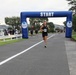 A running race event is underway on a paved road, with a prominent blue inflatable 'START' arch sponsored by MWR (Morale, Welfare and Recreation). A male runner wearing bib number 2350 and a black tank top is the lead runner passing under the arch. Additional runners and spectators are visible in the background, suggesting a military base or organized community race.
