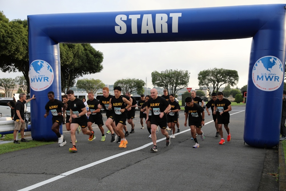 A group of U.S. Army soldiers are crossing the start line of a road race organized under a blue inflatable arch labeled 'START.' The arch displays the U.S. Army MWR (Morale, Welfare, and Recreation) logo on both pillars. Participants are wearing matching black Army PT (Physical Training) uniforms with numbered race bibs.