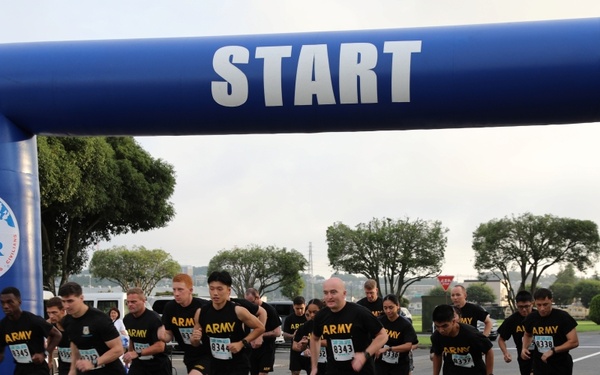 A group of U.S. Army soldiers are crossing the start line of a road race organized under a blue inflatable arch labeled 'START.' The arch displays the U.S. Army MWR (Morale, Welfare, and Recreation) logo on both pillars. Participants are wearing matching black Army PT (Physical Training) uniforms with numbered race bibs.