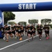 A group of U.S. Army soldiers are crossing the start line of a road race organized under a blue inflatable arch labeled 'START.' The arch displays the U.S. Army MWR (Morale, Welfare, and Recreation) logo on both pillars. Participants are wearing matching black Army PT (Physical Training) uniforms with numbered race bibs.