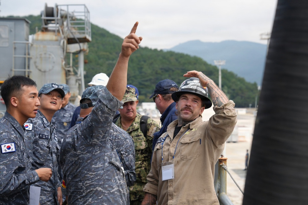 A group of military personnel from multiple nations, including South Korean Navy sailors and U.S. Navy members, are gathered at what appears to be a naval facility or shipyard. A civilian worker wearing a hard hat labeled 'Chief Engineer' and a person in camouflage uniform are pointing upward, likely explaining or directing attention to something overhead. The scene suggests a joint military tour, inspection, or technical briefing at a port or shipyard.