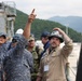 A group of military personnel from multiple nations, including South Korean Navy sailors and U.S. Navy members, are gathered at what appears to be a naval facility or shipyard. A civilian worker wearing a hard hat labeled 'Chief Engineer' and a person in camouflage uniform are pointing upward, likely explaining or directing attention to something overhead. The scene suggests a joint military tour, inspection, or technical briefing at a port or shipyard.