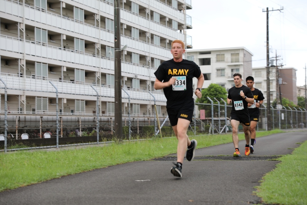 Three runners wearing U.S. Army physical training uniforms (black shirts and shorts) are participating in an organized race event. The lead runner wears bib number 8334, while a visible runner behind him wears bib number 8342. They are running on a paved path alongside a chain-link fence with multi-story buildings visible in the background, suggesting a military installation setting.