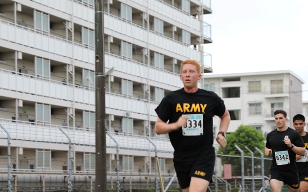 Three runners wearing U.S. Army physical training uniforms (black shirts and shorts) are participating in an organized race event. The lead runner wears bib number 8334, while a visible runner behind him wears bib number 8342. They are running on a paved path alongside a chain-link fence with multi-story buildings visible in the background, suggesting a military installation setting.