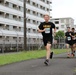 Three runners wearing U.S. Army physical training uniforms (black shirts and shorts) are participating in an organized race event. The lead runner wears bib number 8334, while a visible runner behind him wears bib number 8342. They are running on a paved path alongside a chain-link fence with multi-story buildings visible in the background, suggesting a military installation setting.