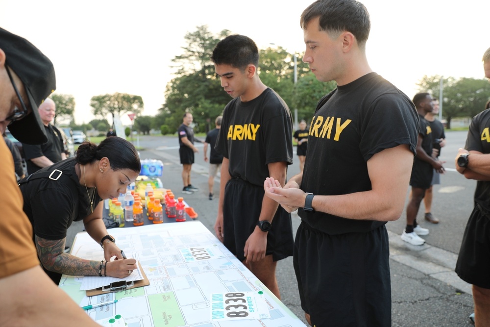 A group of U.S. Army personnel in black PT (physical training) uniforms are gathered at an outdoor registration or check-in table for what appears to be a military run or fitness event. A female soldier with visible tattoos is writing on a clipboard at the table, while two male soldiers stand in front of her holding race bibs numbered 8338. Sports drinks (Gatorade) and water bottles are visible on the table as refreshments.