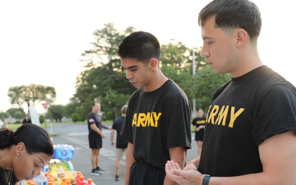 A group of U.S. Army personnel in black PT (physical training) uniforms are gathered at an outdoor registration or check-in table for what appears to be a military run or fitness event. A female soldier with visible tattoos is writing on a clipboard at the table, while two male soldiers stand in front of her holding race bibs numbered 8338. Sports drinks (Gatorade) and water bottles are visible on the table as refreshments.
