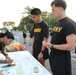 A group of U.S. Army personnel in black PT (physical training) uniforms are gathered at an outdoor registration or check-in table for what appears to be a military run or fitness event. A female soldier with visible tattoos is writing on a clipboard at the table, while two male soldiers stand in front of her holding race bibs numbered 8338. Sports drinks (Gatorade) and water bottles are visible on the table as refreshments.