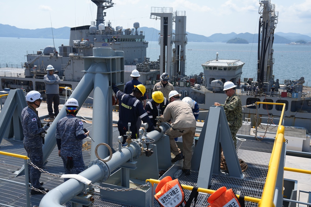 The image shows a multinational naval replenishment-at-sea or pier-side fueling operation involving personnel from what appears to be the Republic of Korea Navy and the U.S. Navy, along with civilian contractors. Personnel are working on fuel transfer piping and valve connections on the deck of a naval vessel. Multiple ships are visible in the background in a harbor or anchorage setting.