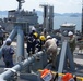 The image shows a multinational naval replenishment-at-sea or pier-side fueling operation involving personnel from what appears to be the Republic of Korea Navy and the U.S. Navy, along with civilian contractors. Personnel are working on fuel transfer piping and valve connections on the deck of a naval vessel. Multiple ships are visible in the background in a harbor or anchorage setting.