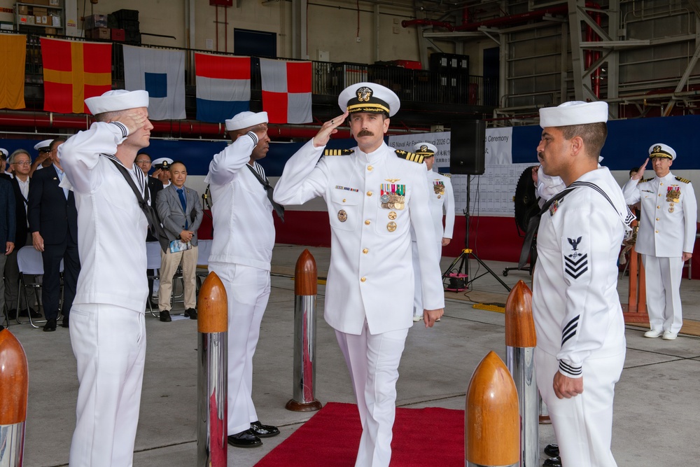 A formal U.S. Navy change of command or commissioning ceremony is taking place inside a military hangar. A senior officer in dress whites with gold insignia and medals is rendering a salute while enlisted sailors in white uniforms salute in return. Naval signal flags are displayed in the background alongside a ceremony banner, and civilian observers are seated to the left.