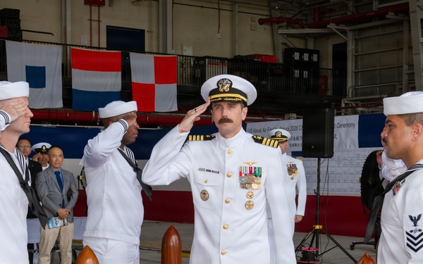 A formal U.S. Navy change of command or commissioning ceremony is taking place inside a military hangar. A senior officer in dress whites with gold insignia and medals is rendering a salute while enlisted sailors in white uniforms salute in return. Naval signal flags are displayed in the background alongside a ceremony banner, and civilian observers are seated to the left.