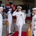 A formal U.S. Navy change of command or commissioning ceremony is taking place inside a military hangar. A senior officer in dress whites with gold insignia and medals is rendering a salute while enlisted sailors in white uniforms salute in return. Naval signal flags are displayed in the background alongside a ceremony banner, and civilian observers are seated to the left.