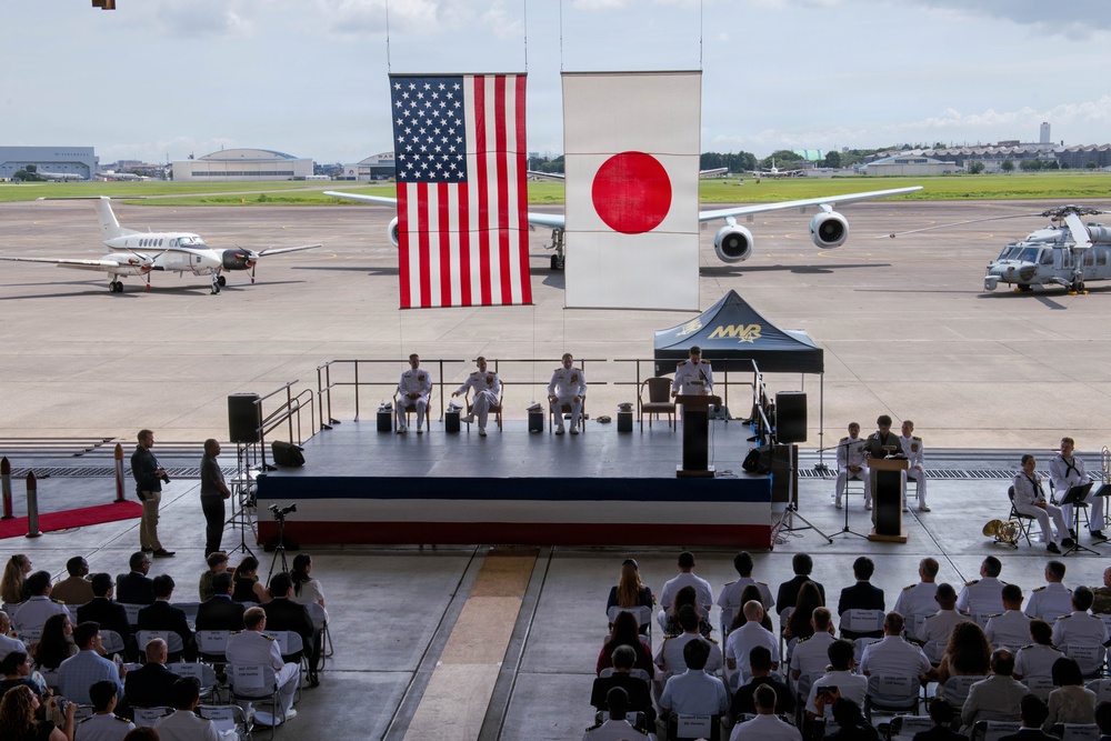 A formal military ceremony is taking place on an outdoor stage at what appears to be a joint U.S.-Japanese military airbase. Large American and Japanese flags are displayed prominently as backdrops, with military personnel in white dress uniforms seated on stage and in the audience. Aircraft are visible on the tarmac in the background, and a military band is positioned to the right of the stage.
