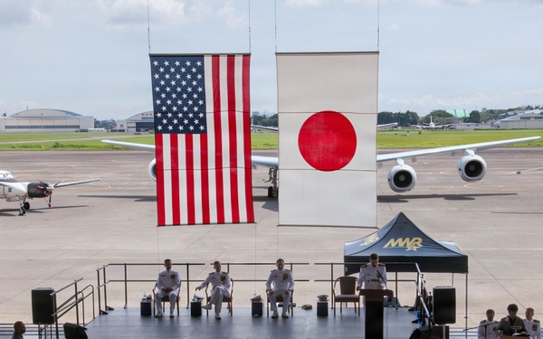 A formal military ceremony is taking place on an outdoor stage at what appears to be a joint U.S.-Japanese military airbase. Large American and Japanese flags are displayed prominently as backdrops, with military personnel in white dress uniforms seated on stage and in the audience. Aircraft are visible on the tarmac in the background, and a military band is positioned to the right of the stage.