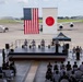 A formal military ceremony is taking place on an outdoor stage at what appears to be a joint U.S.-Japanese military airbase. Large American and Japanese flags are displayed prominently as backdrops, with military personnel in white dress uniforms seated on stage and in the audience. Aircraft are visible on the tarmac in the background, and a military band is positioned to the right of the stage.