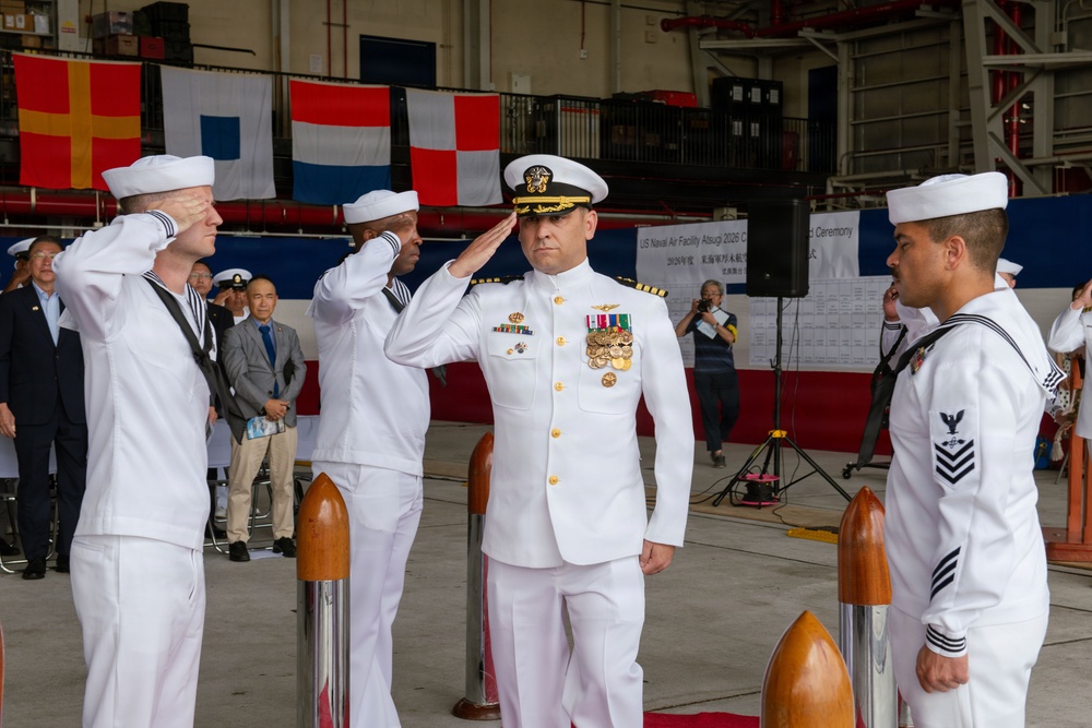 A formal U.S. Navy military ceremony is taking place inside a hangar facility, identified by a partially visible sign as 'US Naval Air Facility Atsugi 2026 Change of Command Ceremony.' A senior officer in white dress uniform with gold insignia and multiple medal ribbons is rendering a salute, flanked by enlisted sailors also saluting. Civilian and military observers are visible in the background alongside international naval signal flags.