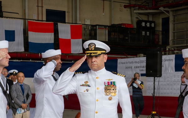 A formal U.S. Navy military ceremony is taking place inside a hangar facility, identified by a partially visible sign as 'US Naval Air Facility Atsugi 2026 Change of Command Ceremony.' A senior officer in white dress uniform with gold insignia and multiple medal ribbons is rendering a salute, flanked by enlisted sailors also saluting. Civilian and military observers are visible in the background alongside international naval signal flags.