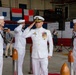 A formal U.S. Navy military ceremony is taking place inside a hangar facility, identified by a partially visible sign as 'US Naval Air Facility Atsugi 2026 Change of Command Ceremony.' A senior officer in white dress uniform with gold insignia and multiple medal ribbons is rendering a salute, flanked by enlisted sailors also saluting. Civilian and military observers are visible in the background alongside international naval signal flags.