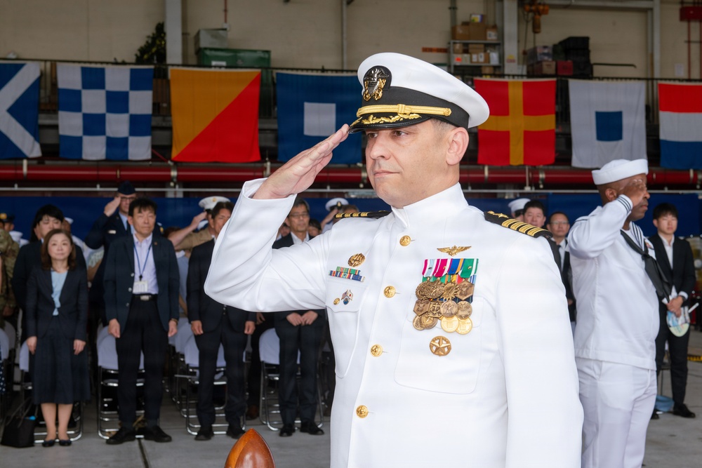 A senior U.S. Navy officer in dress white uniform is rendering a formal salute during what appears to be a military ceremony. The officer displays numerous medals, ribbons, and insignia consistent with a high-ranking captain or flag officer. The event appears to be held in a hangar or large indoor facility decorated with naval signal flags, with a mixed civilian and military audience in the background.