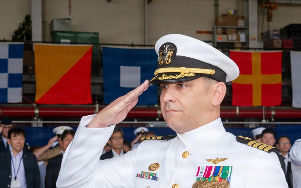 A senior U.S. Navy officer in dress white uniform is rendering a formal salute during what appears to be a military ceremony. The officer displays numerous medals, ribbons, and insignia consistent with a high-ranking captain or flag officer. The event appears to be held in a hangar or large indoor facility decorated with naval signal flags, with a mixed civilian and military audience in the background.