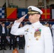 A senior U.S. Navy officer in dress white uniform is rendering a formal salute during what appears to be a military ceremony. The officer displays numerous medals, ribbons, and insignia consistent with a high-ranking captain or flag officer. The event appears to be held in a hangar or large indoor facility decorated with naval signal flags, with a mixed civilian and military audience in the background.