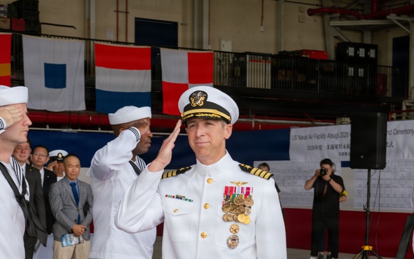 A formal U.S. Navy change of command or award ceremony is taking place inside a hangar at US Naval Air Facility Atsugi, Japan. A senior officer in dress whites with extensive medal decorations and gold cap insignia is rendering a salute alongside enlisted sailors in white uniforms. Civilian dignitaries and military personnel are visible in the background observing the ceremony.