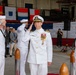 A formal U.S. Navy change of command or award ceremony is taking place inside a hangar at US Naval Air Facility Atsugi, Japan. A senior officer in dress whites with extensive medal decorations and gold cap insignia is rendering a salute alongside enlisted sailors in white uniforms. Civilian dignitaries and military personnel are visible in the background observing the ceremony.