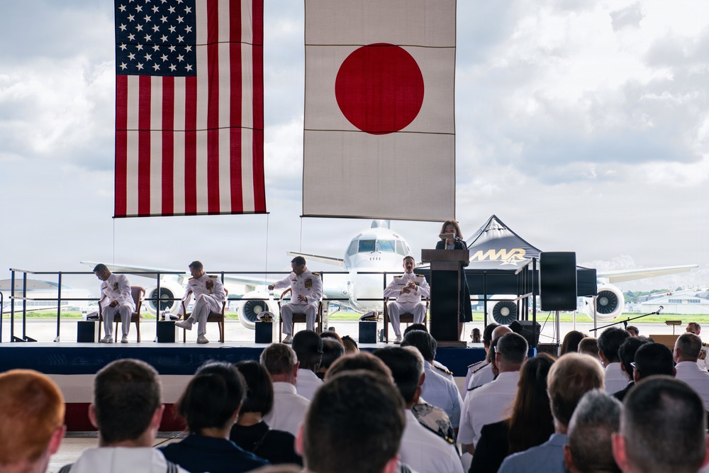 A formal military ceremony is taking place outdoors on an airfield, with both the United States and Japanese flags prominently displayed. Four naval officers in white dress uniforms are seated on a raised stage while a civilian woman speaks at a podium. A large military aircraft is visible in the background, and a sizeable audience of military personnel and civilians is seated facing the stage.