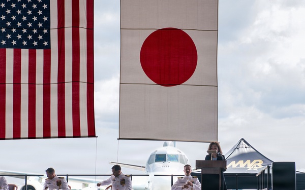 A formal military ceremony is taking place outdoors on an airfield, with both the United States and Japanese flags prominently displayed. Four naval officers in white dress uniforms are seated on a raised stage while a civilian woman speaks at a podium. A large military aircraft is visible in the background, and a sizeable audience of military personnel and civilians is seated facing the stage.