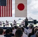 A formal military ceremony is taking place outdoors on an airfield, with both the United States and Japanese flags prominently displayed. Four naval officers in white dress uniforms are seated on a raised stage while a civilian woman speaks at a podium. A large military aircraft is visible in the background, and a sizeable audience of military personnel and civilians is seated facing the stage.