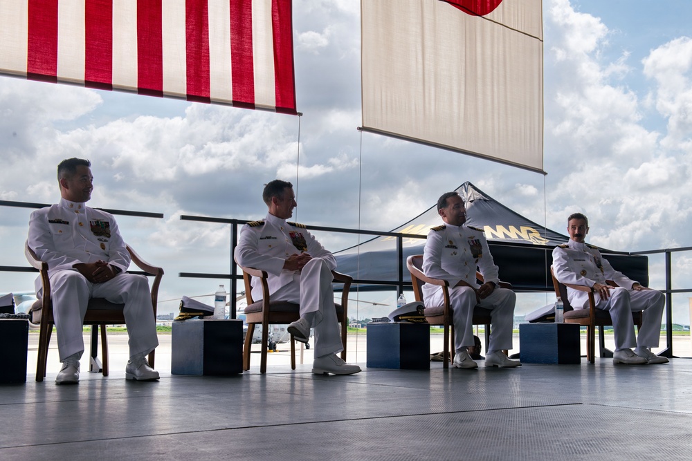 Four naval officers in white dress uniforms are seated on a raised platform or stage during what appears to be a formal military ceremony. Two flags are visible overhead, including what appears to be the United States flag and a Japanese flag, suggesting a bilateral or joint military event. The setting appears to be an outdoor or semi-open hangar area at a military airfield.