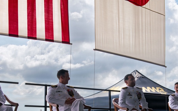 Four naval officers in white dress uniforms are seated on a raised platform or stage during what appears to be a formal military ceremony. Two flags are visible overhead, including what appears to be the United States flag and a Japanese flag, suggesting a bilateral or joint military event. The setting appears to be an outdoor or semi-open hangar area at a military airfield.