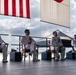 Four naval officers in white dress uniforms are seated on a raised platform or stage during what appears to be a formal military ceremony. Two flags are visible overhead, including what appears to be the United States flag and a Japanese flag, suggesting a bilateral or joint military event. The setting appears to be an outdoor or semi-open hangar area at a military airfield.
