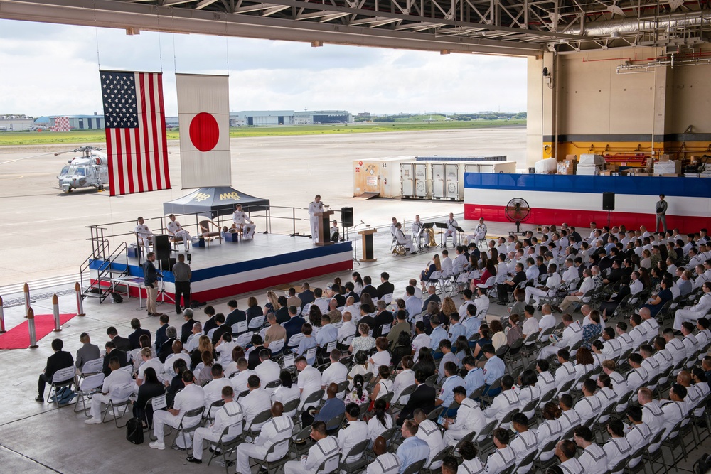 A formal military ceremony is taking place inside a large aircraft hangar, with hundreds of attendees seated in folding chairs facing a decorated stage. Both the American and Japanese flags are prominently displayed, indicating a joint U.S.-Japan military event, likely a change of command or commissioning ceremony. Military personnel in white dress uniforms make up the majority of the audience, with civilians interspersed throughout.