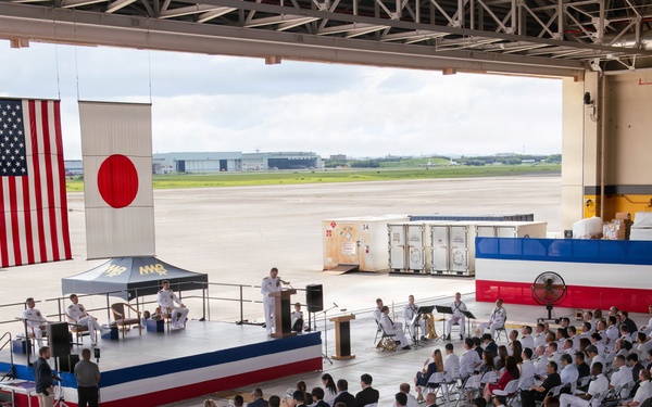 A formal military ceremony is taking place inside a large aircraft hangar, with hundreds of attendees seated in folding chairs facing a decorated stage. Both the American and Japanese flags are prominently displayed, indicating a joint U.S.-Japan military event, likely a change of command or commissioning ceremony. Military personnel in white dress uniforms make up the majority of the audience, with civilians interspersed throughout.