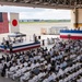 A formal military ceremony is taking place inside a large aircraft hangar, with hundreds of attendees seated in folding chairs facing a decorated stage. Both the American and Japanese flags are prominently displayed, indicating a joint U.S.-Japan military event, likely a change of command or commissioning ceremony. Military personnel in white dress uniforms make up the majority of the audience, with civilians interspersed throughout.