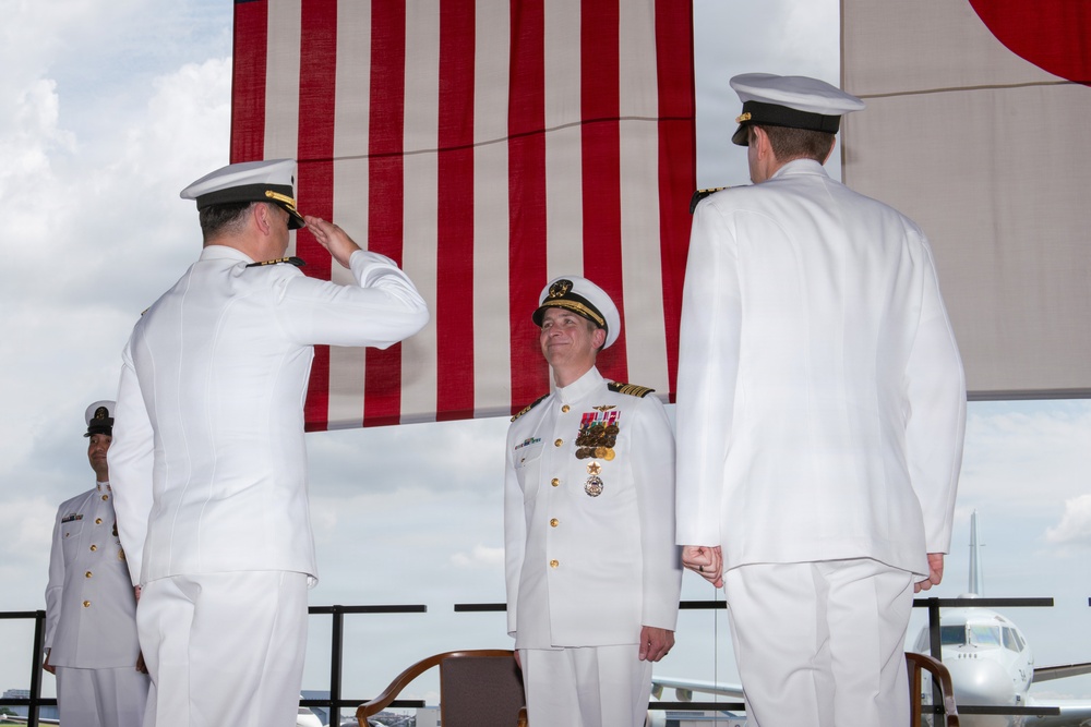 A formal U.S. Navy military ceremony is taking place outdoors, likely a change of command or retirement ceremony. Three naval officers in white dress uniforms are prominently featured, with one officer rendering a salute to a centrally positioned officer wearing multiple medals and ribbons. A large American flag serves as the backdrop.