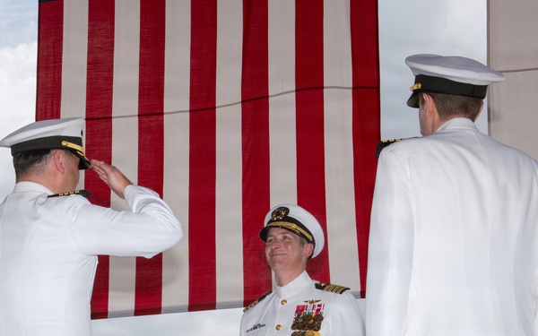 A formal U.S. Navy military ceremony is taking place outdoors, likely a change of command or retirement ceremony. Three naval officers in white dress uniforms are prominently featured, with one officer rendering a salute to a centrally positioned officer wearing multiple medals and ribbons. A large American flag serves as the backdrop.