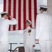 A formal U.S. Navy military ceremony is taking place outdoors, likely a change of command or retirement ceremony. Three naval officers in white dress uniforms are prominently featured, with one officer rendering a salute to a centrally positioned officer wearing multiple medals and ribbons. A large American flag serves as the backdrop.
