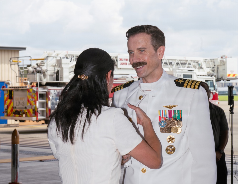 A naval officer in white dress uniform is being decorated or having insignia pinned by a woman in a white outfit, suggesting a military promotion or award ceremony. The officer displays numerous medals, ribbons, and command insignia on his uniform, indicating significant service and rank. The event appears to be taking place outdoors on a tarmac or airfield, with fire trucks visible in the background.