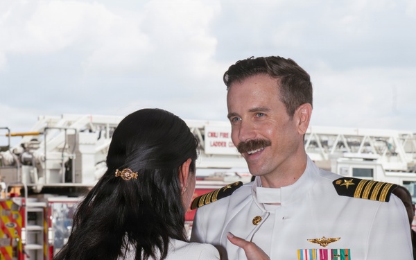 A naval officer in white dress uniform is being decorated or having insignia pinned by a woman in a white outfit, suggesting a military promotion or award ceremony. The officer displays numerous medals, ribbons, and command insignia on his uniform, indicating significant service and rank. The event appears to be taking place outdoors on a tarmac or airfield, with fire trucks visible in the background.