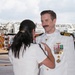 A naval officer in white dress uniform is being decorated or having insignia pinned by a woman in a white outfit, suggesting a military promotion or award ceremony. The officer displays numerous medals, ribbons, and command insignia on his uniform, indicating significant service and rank. The event appears to be taking place outdoors on a tarmac or airfield, with fire trucks visible in the background.
